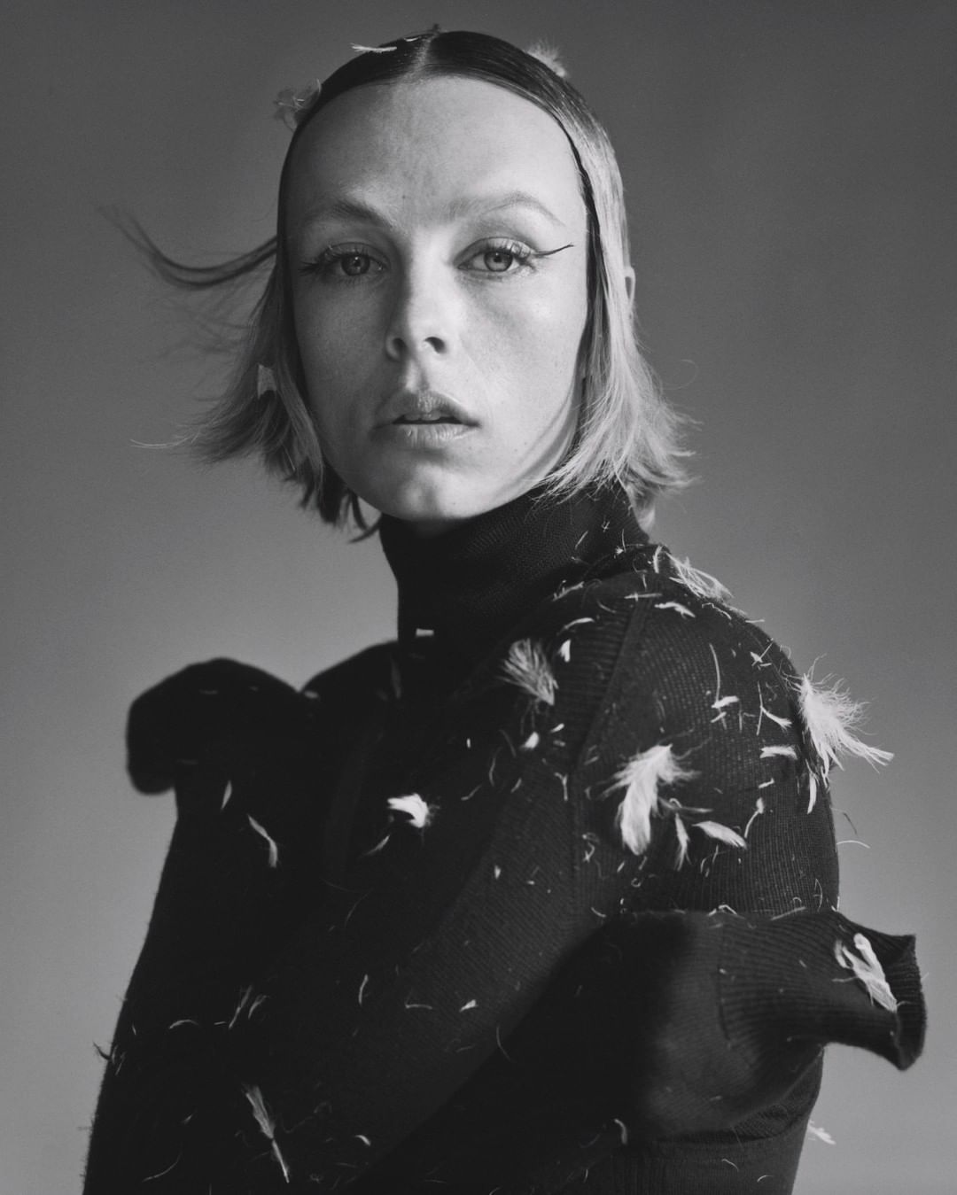 Portrait of a blonde woman with dark eyeliner wearing a black turtleneck sweater with white feathers in a studio with grey background.