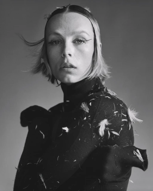Portrait of a blonde woman with dark eyeliner wearing a black turtleneck sweater with white feathers in a studio with grey background.