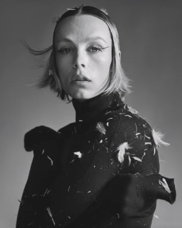Portrait of a blonde woman with dark eyeliner wearing a black turtleneck sweater with white feathers in a studio with grey background.