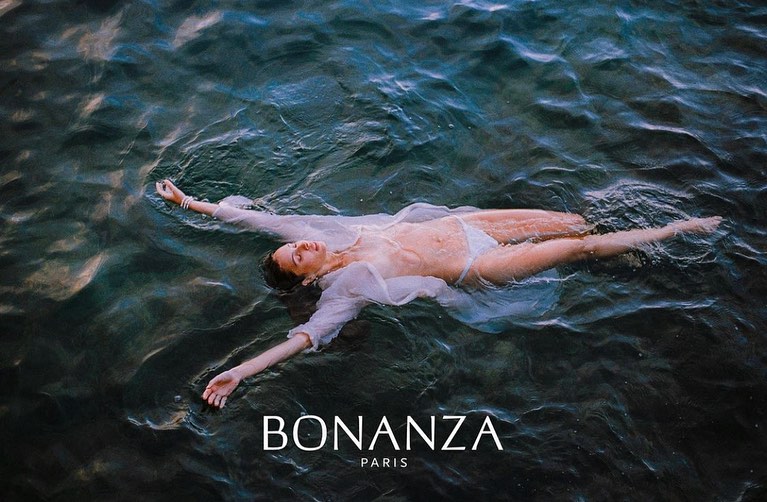 Woman in white swimwear and top lying on her back in a body of water with visible waves, outdoor setting. Brand logo in the corner.