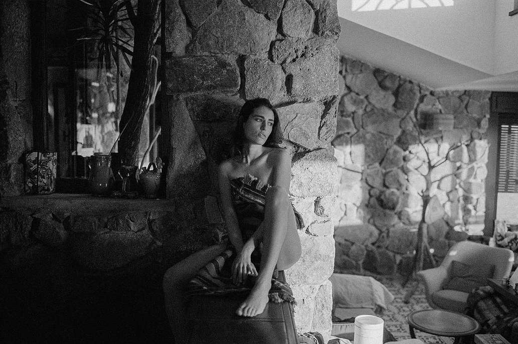 Black and white portrait of a slender adult woman posing topless while sitting against a stone fireplace in a rustic home interior.
