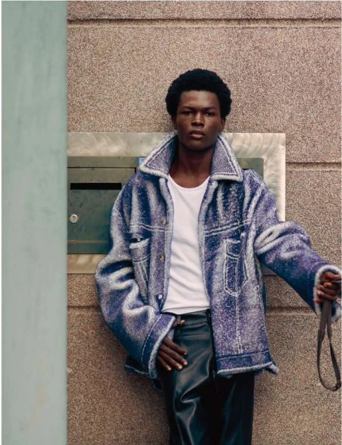 Eye-level shot of African-American male model wearing a denim sherpa jacket and leather pants. He poses against a textured wall.
