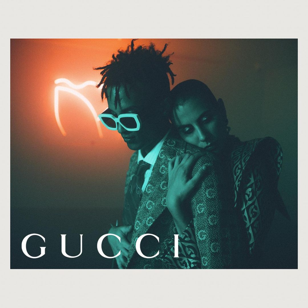 Fashion-forward duo in Gucci apparel: a stylish man with shades and a patterned blazer, with a woman embracing his shoulder.