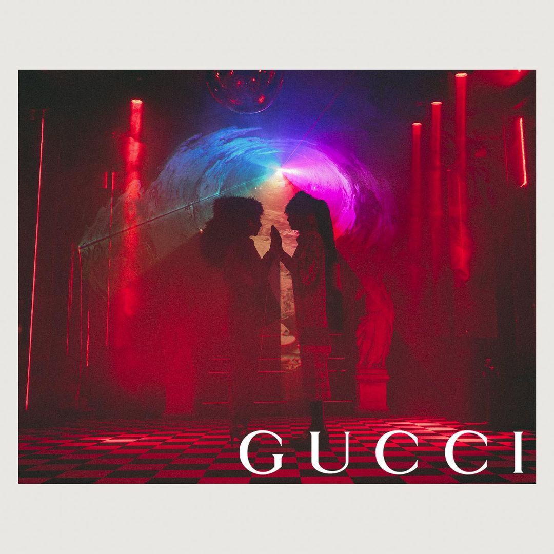 Two people silhouetted against colorful light display with disco ball and Gucci logo on a checkered floor, red and blue tones.