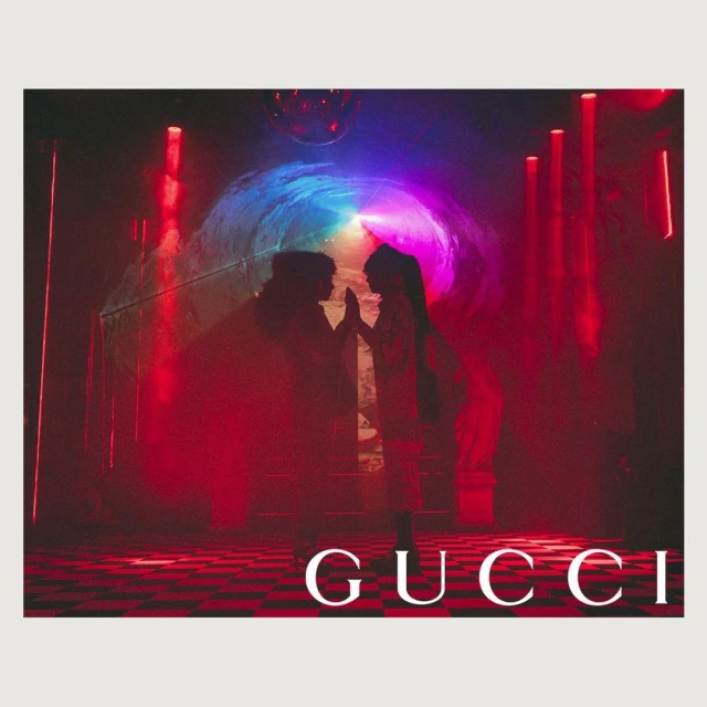 Two people silhouetted against colorful light display with disco ball and Gucci logo on a checkered floor, red and blue tones.