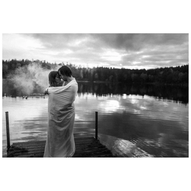 Black and white photo of a couple wrapped in a blanket embracing on a wooden dock with a lake and forest in the background.