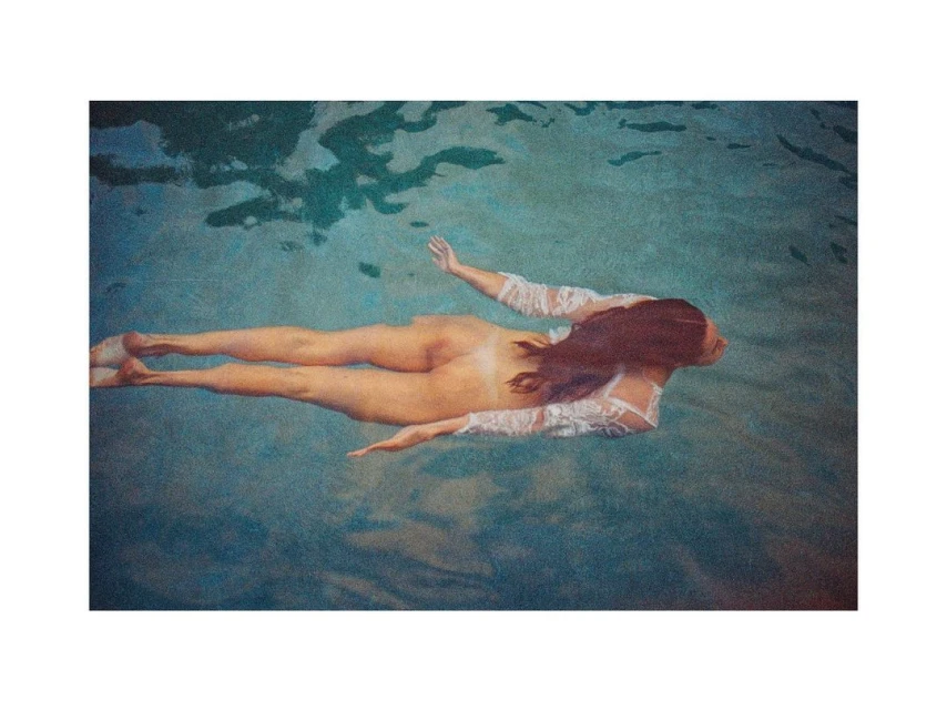 A woman with long brown hair and a long-sleeved white lace top floats horizontally in teal-blue water. The scene has an artful, grainy aesthetic.