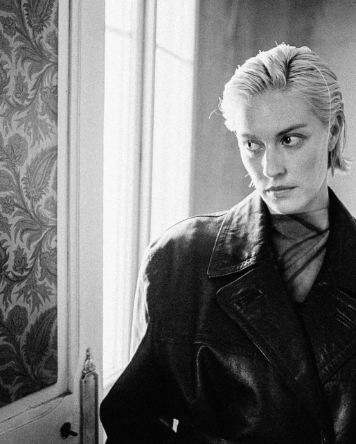 Black and white portrait of an adult female with short blonde hair wearing a dark leather jacket standing indoors by a window.