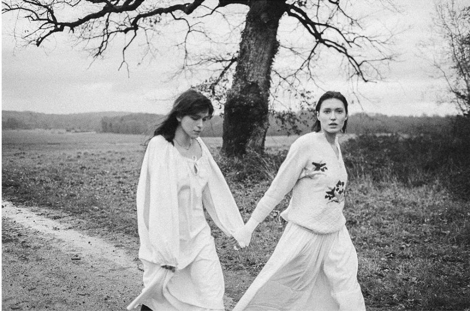 Two women in vintage dresses walk hand-in-hand on a path in a rural landscape under a cloudy sky. Black and white photograph.