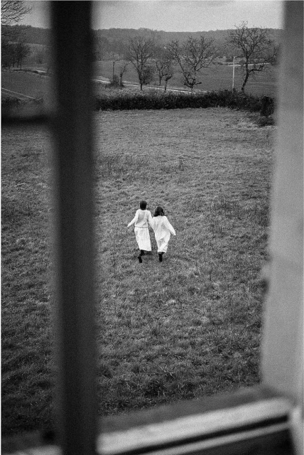 Black and white photograph of two figures in white coats walking in a grassy field, as viewed through a window frame. Rural scene.