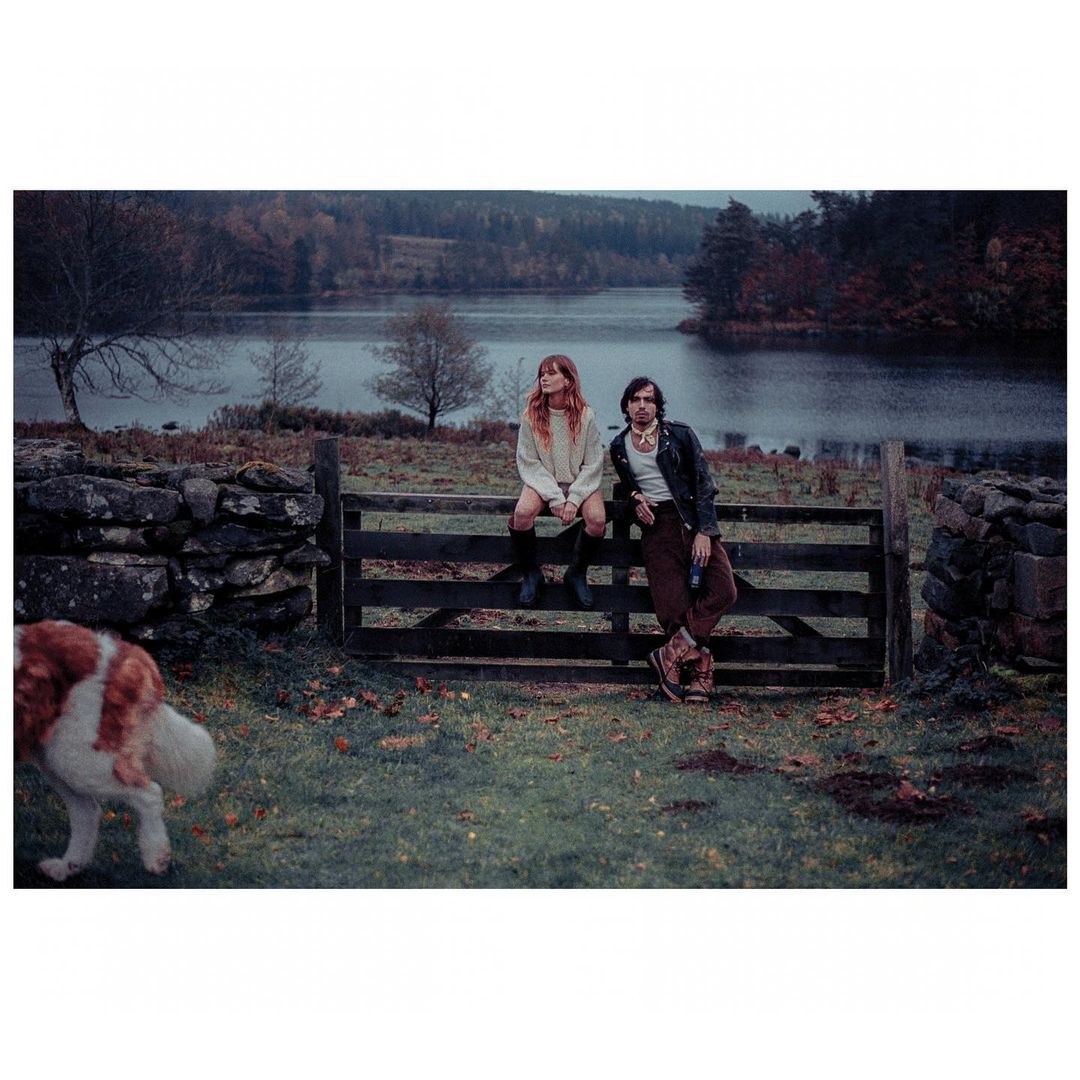 A couple sits on a wooden fence in a rural field by a lake, with an approaching dog, wearing casual clothing and looking toward the camera.