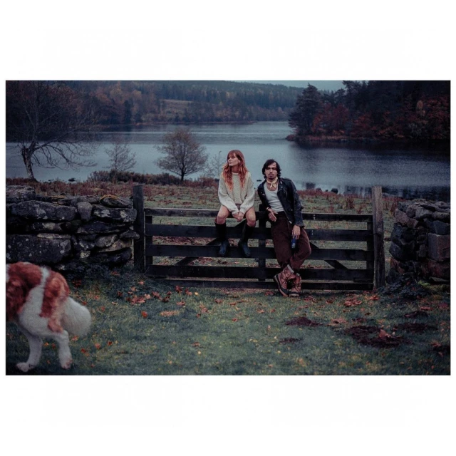 A couple sits on a wooden fence in a rural field by a lake, with an approaching dog, wearing casual clothing and looking toward the camera.