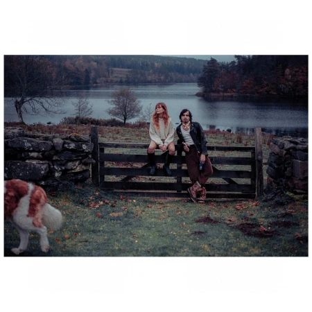 A couple sits on a wooden fence in a rural field by a lake, with an approaching dog, wearing casual clothing and looking toward the camera.