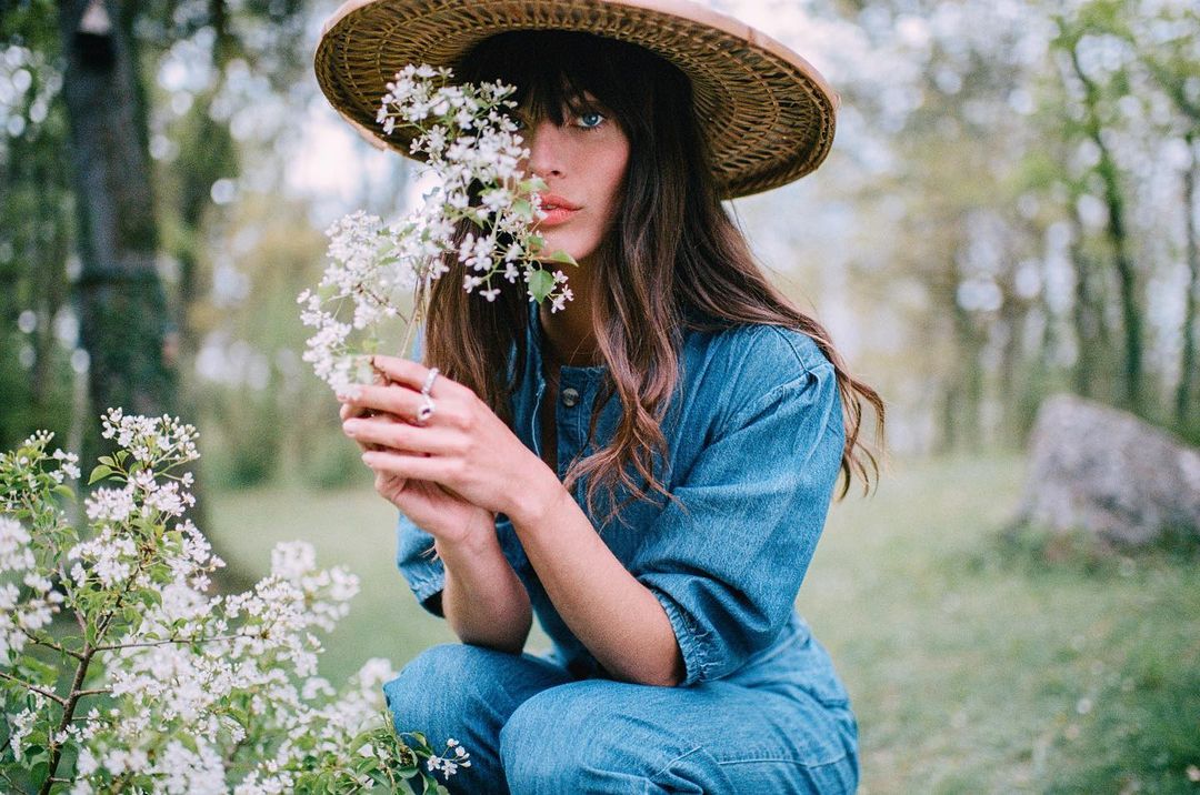 Young woman in a blue denim jumpsuit and straw hat sits in a field of white flowers in a garden setting, holding flowers.