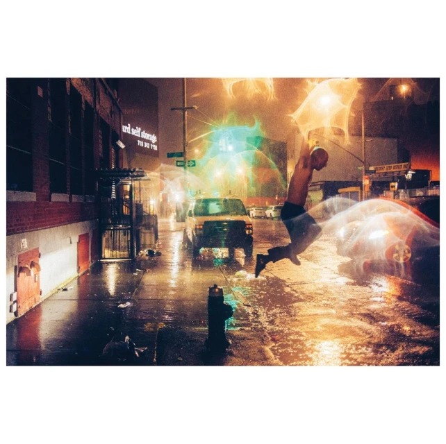 Man jumps on city street at night surrounded by cars. Building with Unit Self Storage sign visible. Street is wet and reflects light.