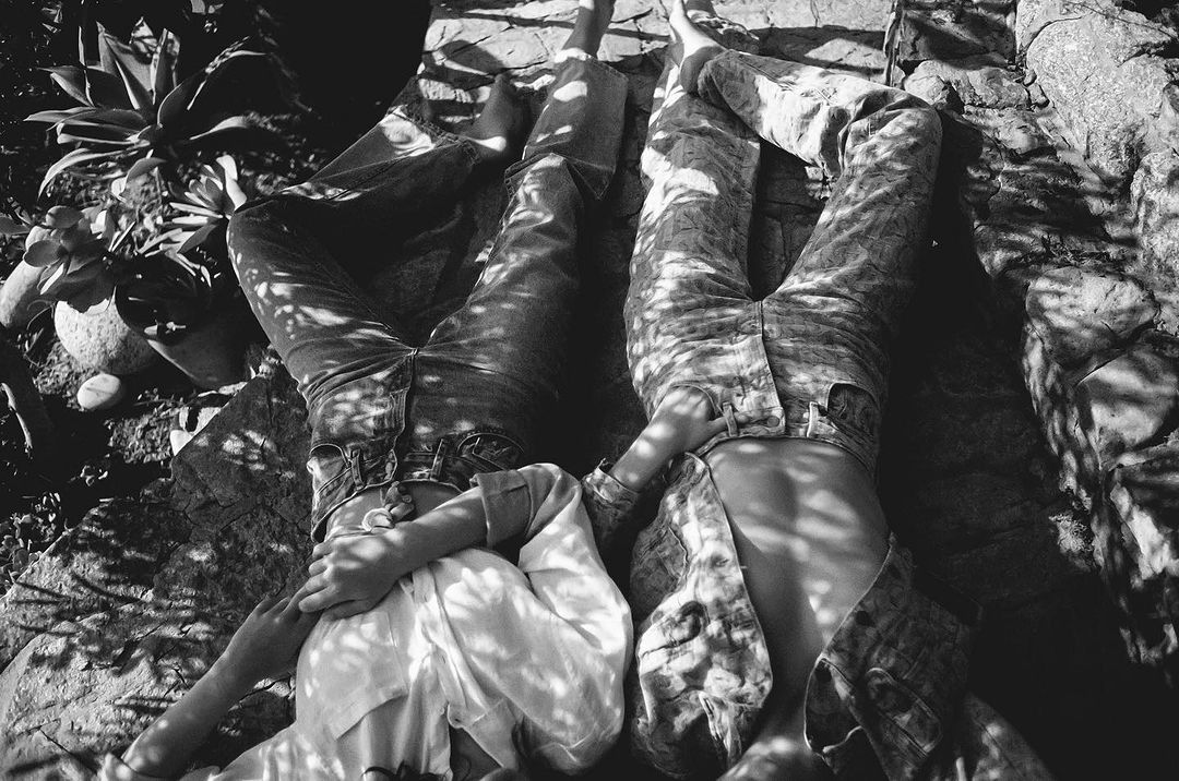 Monochrome close-up of two women lying on rock in jeans and shirts, shadows creating abstract patterns, natural light, relaxed pose.