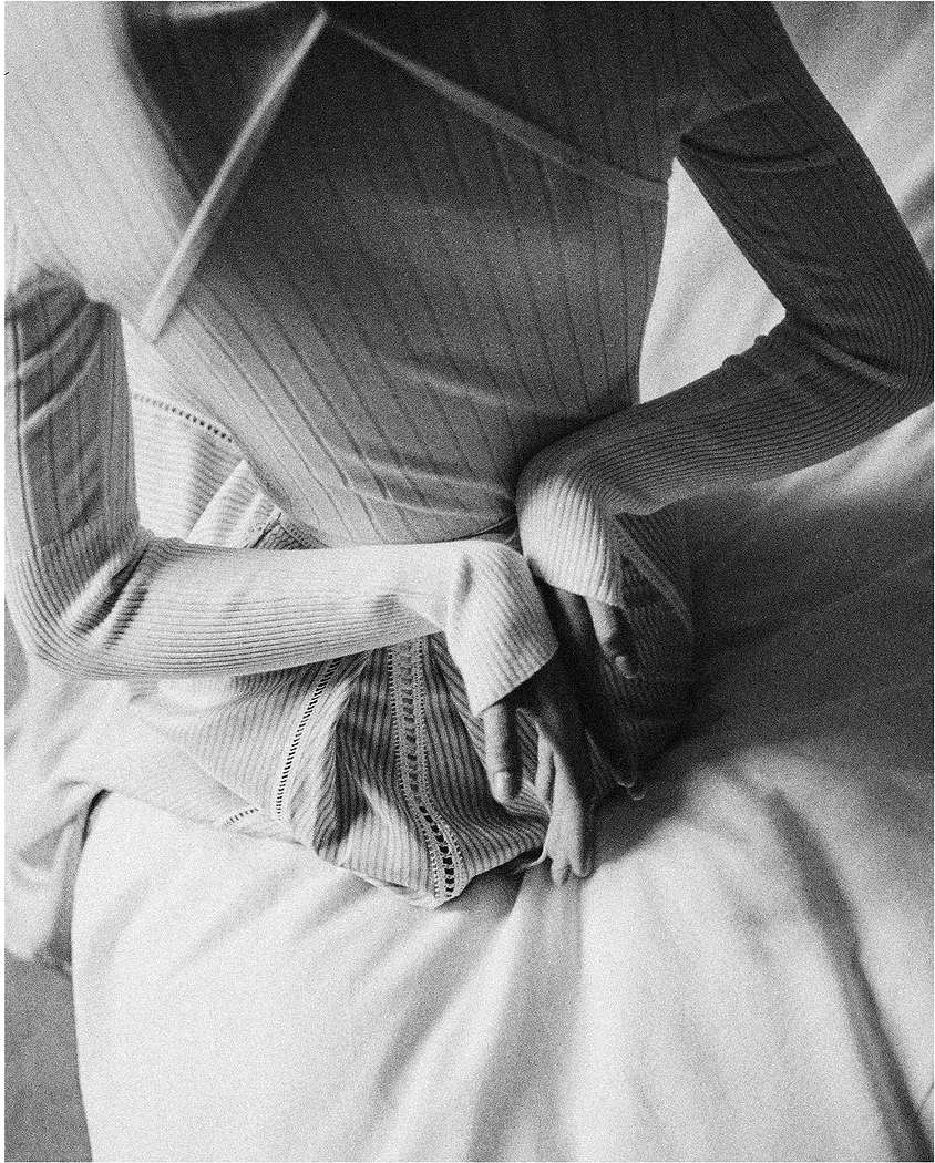 Close-up shot of a woman wearing a long-sleeved top with her hands on her back, while sitting on a white bed in an indoor setting.