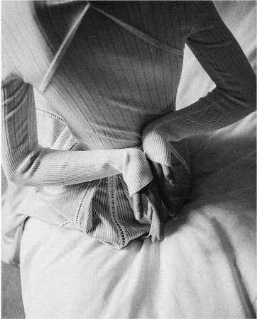 Close-up shot of a woman wearing a long-sleeved top with her hands on her back, while sitting on a white bed in an indoor setting.