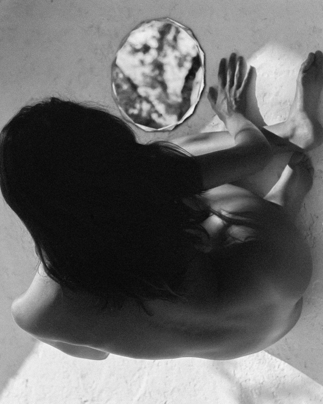 Black and white artistic portrait of a nude adult female seated looking at a round mirror reflecting back the dark cloudy sky.