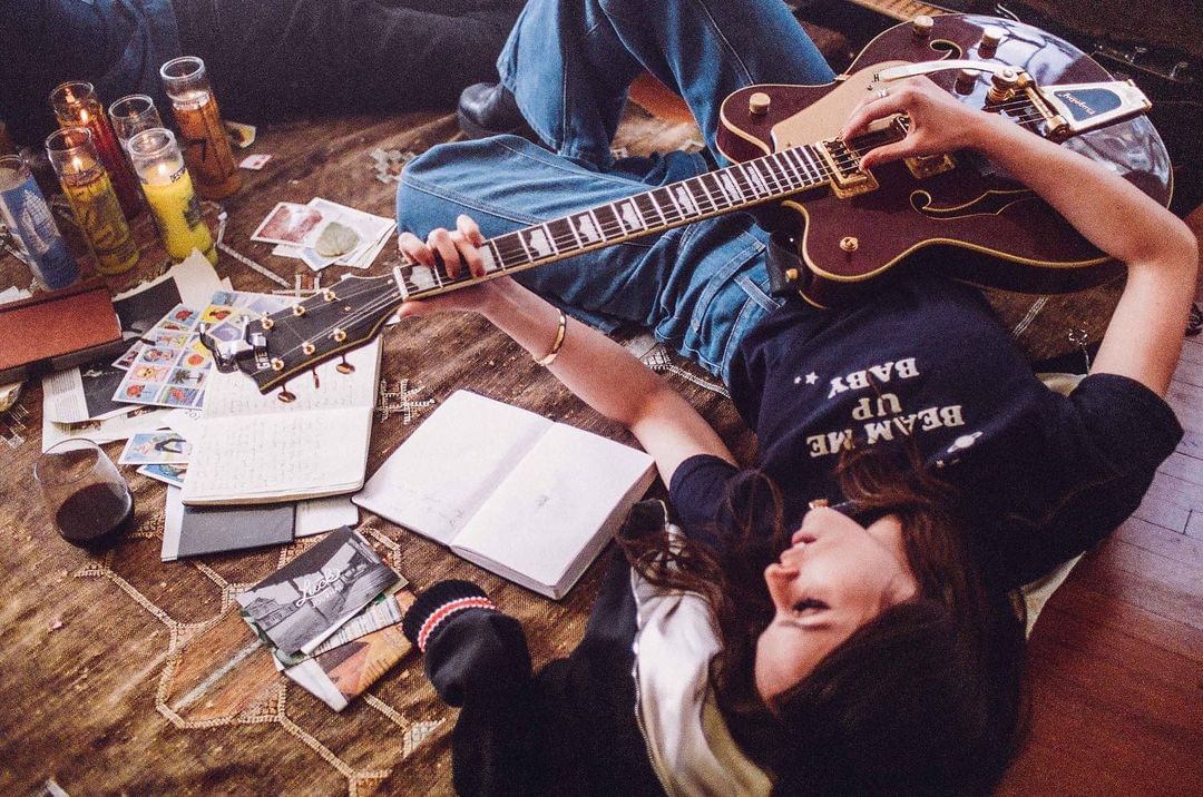 Woman lies on rug surrounded by notebooks, guitar, cards, and candles, casually playing guitar, indoor living room, home interior.