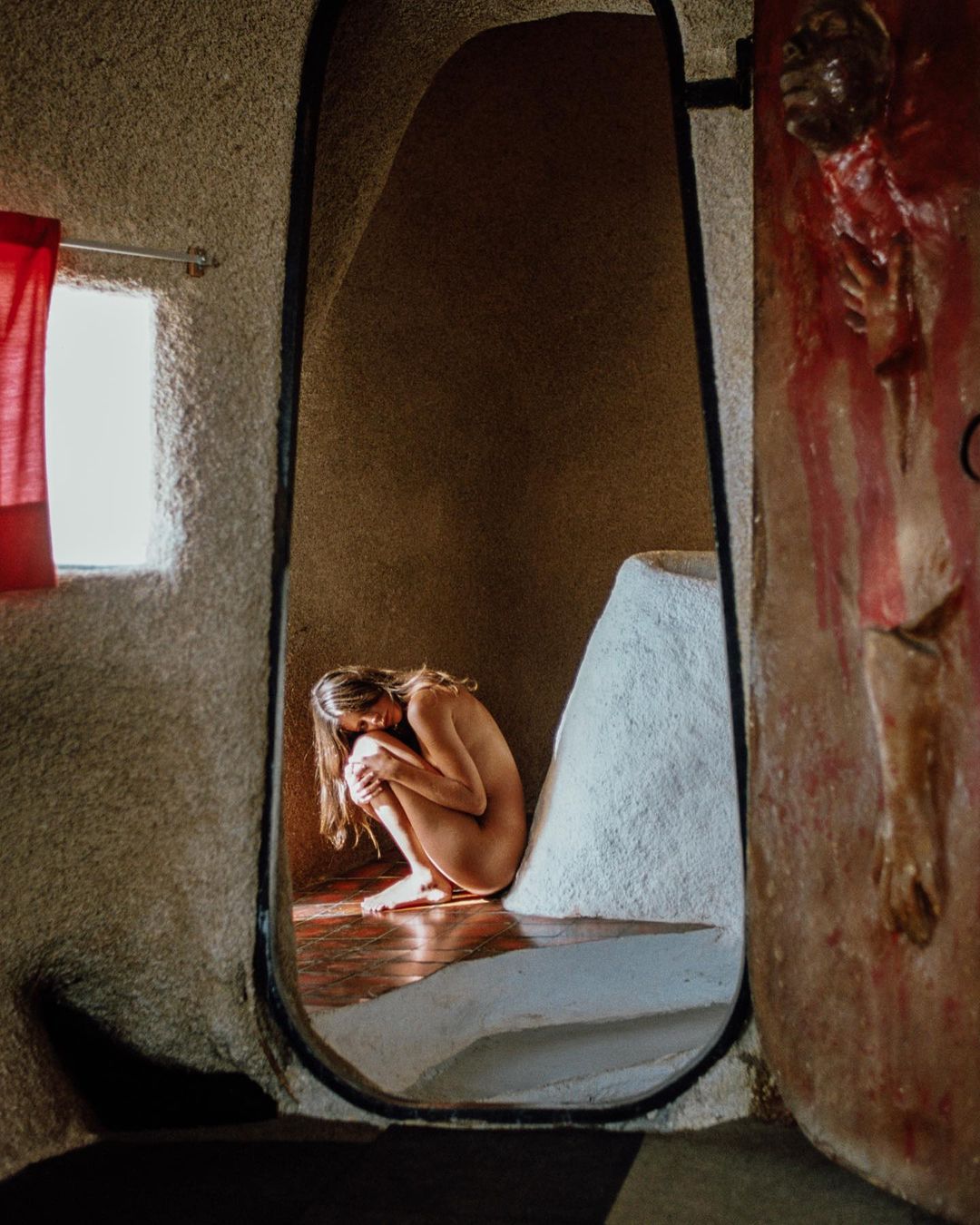 Nude woman with long hair in fetal position inside stone room with cave-like opening, red curtain, and sunlight on wood floor.
