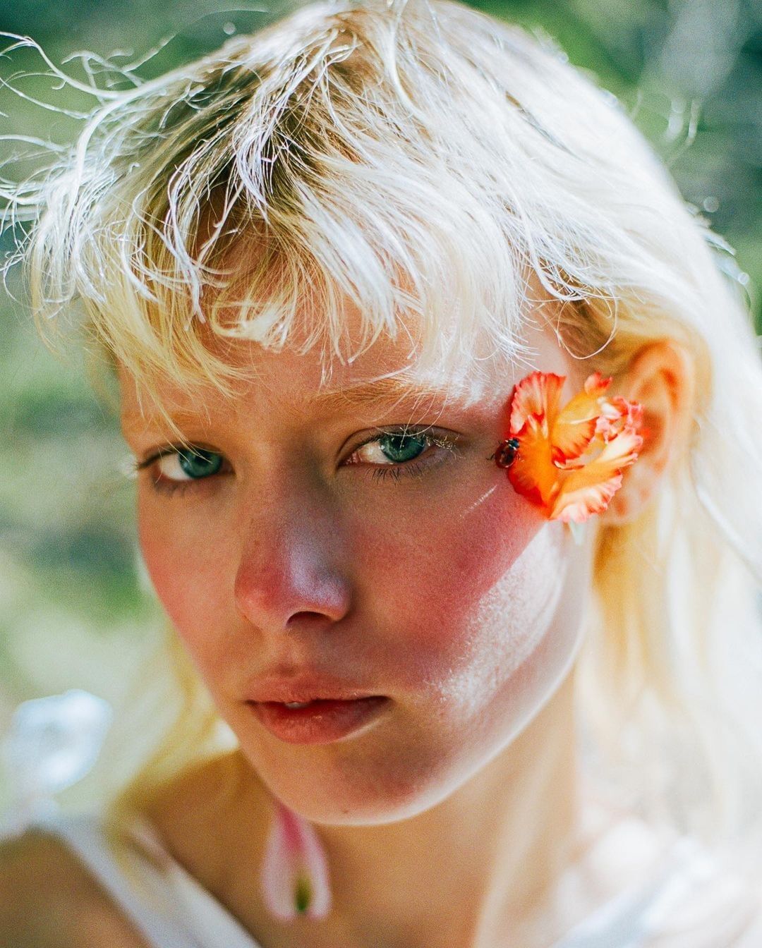Close-up portrait of a young adult woman with blond hair and flower in her hair, captured with a shallow depth of field in outdoor setting.