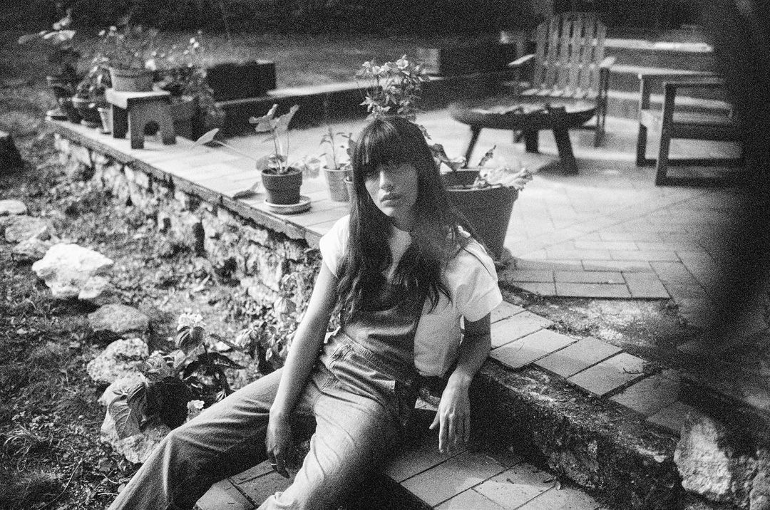 Monochrome portrait of a young woman sitting on a stone ledge outdoors surrounded by potted plants and greenery in a backyard setting.