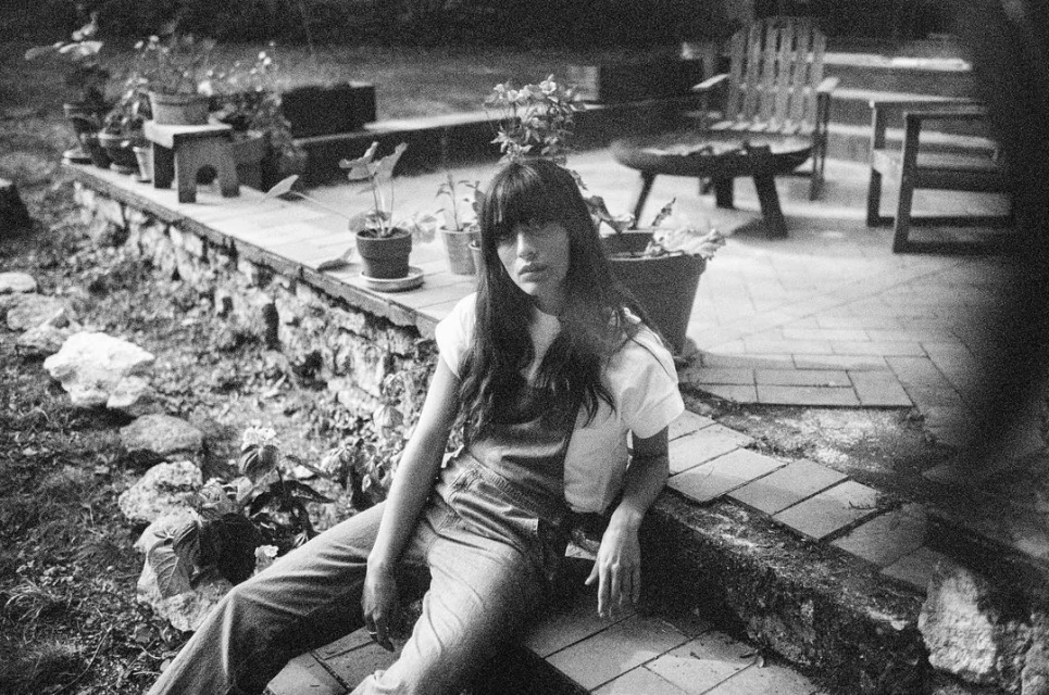 Monochrome portrait of a young woman sitting on a stone ledge outdoors surrounded by potted plants and greenery in a backyard setting.