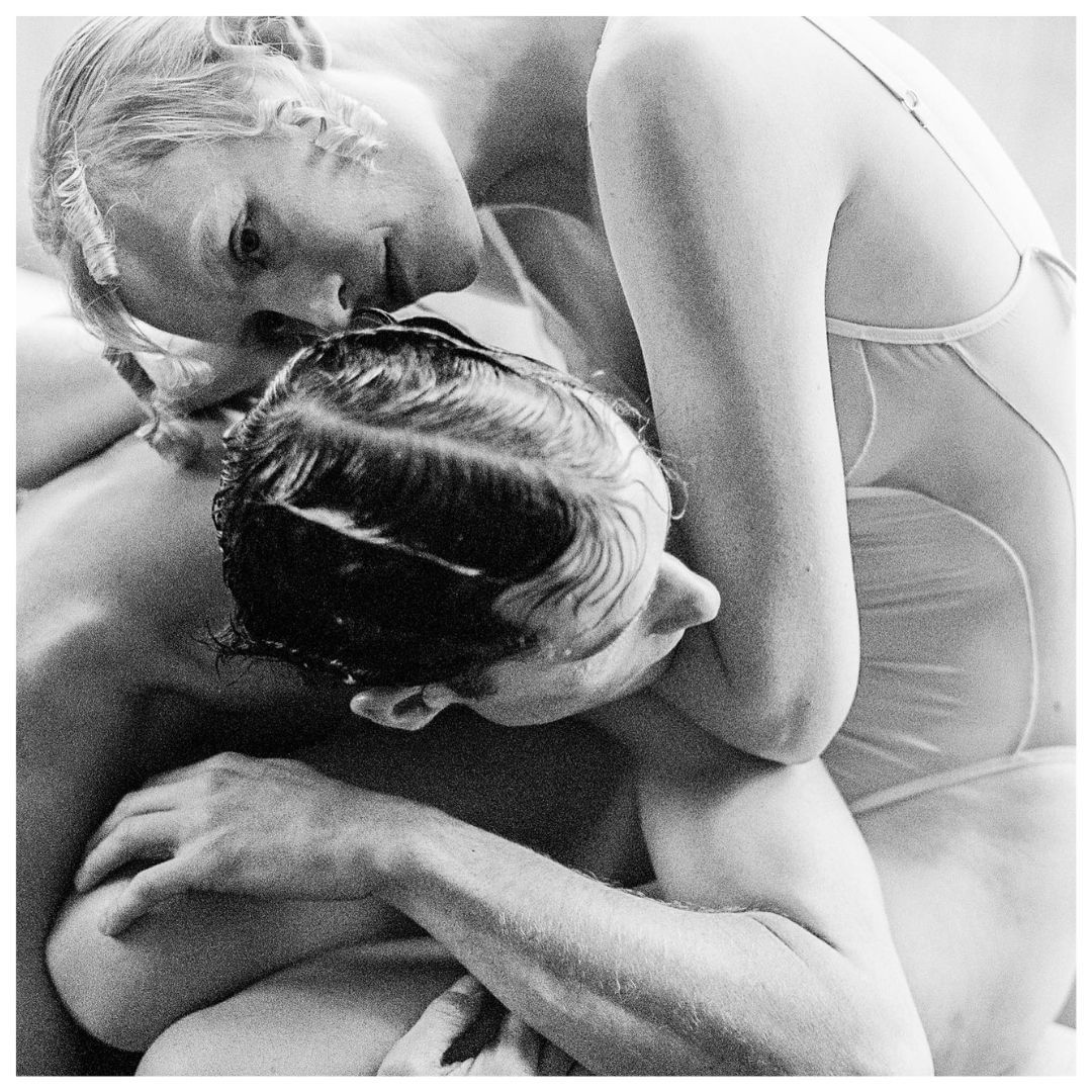 Black-and-white photo of a blond woman in a sheer bodysuit embracing a brunette man with his back to the camera in an indoor setting.