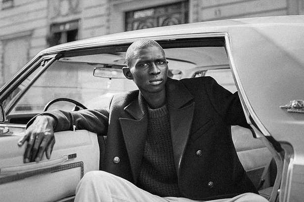 Black and white portrait of a man in a car, wearing a coat and sweater. Urban setting with building in the background.