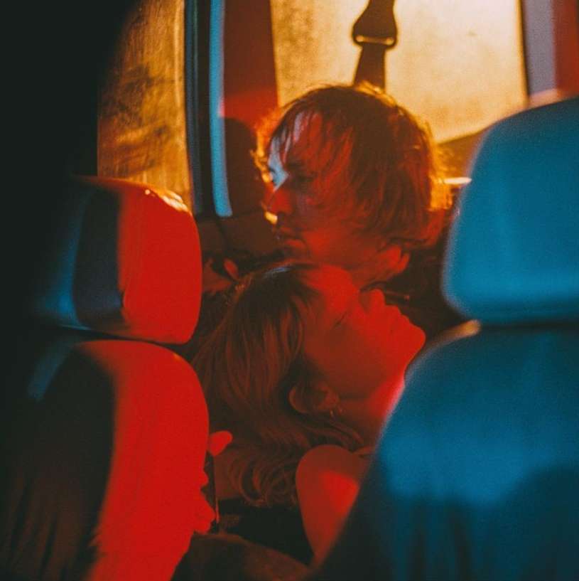 Close-up photo of a couple in an automotive interior, possibly a van, illuminated by red and orange lighting, creating a warm and intimate ambiance.