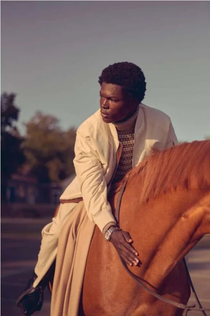 An adult male rides a chestnut horse in an outdoor setting. The man wears a light-colored jacket and trousers, and the horse is saddled.