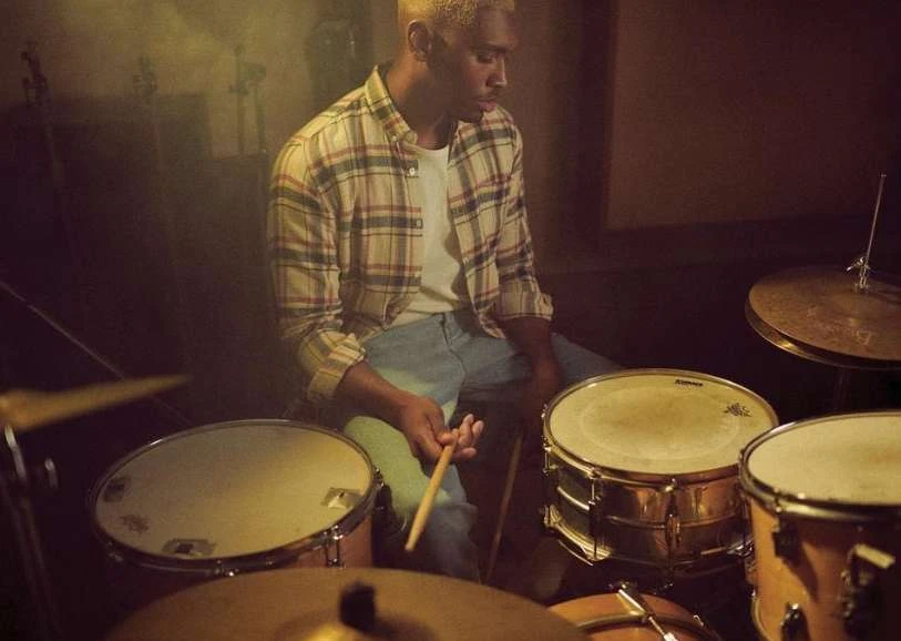 Man playing drums in a music studio. He is holding drumsticks, wearing a plaid shirt, and playing a drum set. The setting is dark and moody.