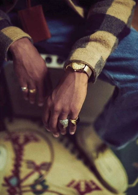 Close-up of a person with tan skin wearing blue jeans and a checked jacket, adorned with rings and a wristwatch, with a vintage amplifier.