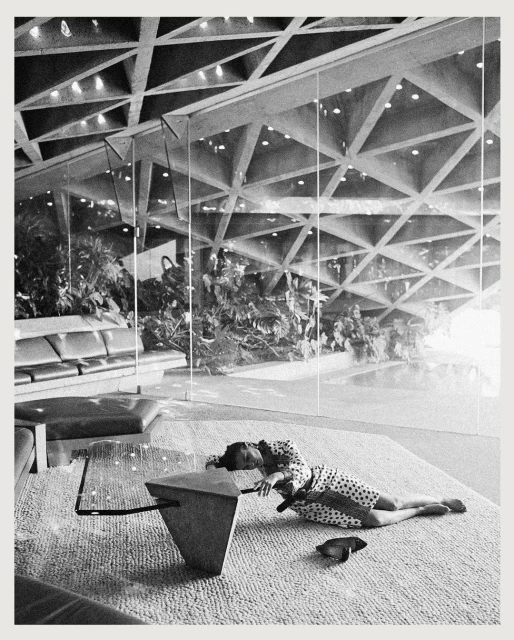 Black and white image features a woman in a polka dot dress lying on a carpet in a modern, architectural interior with a geometric ceiling.