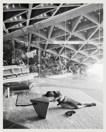Black and white image features a woman in a polka dot dress lying on a carpet in a modern, architectural interior with a geometric ceiling.