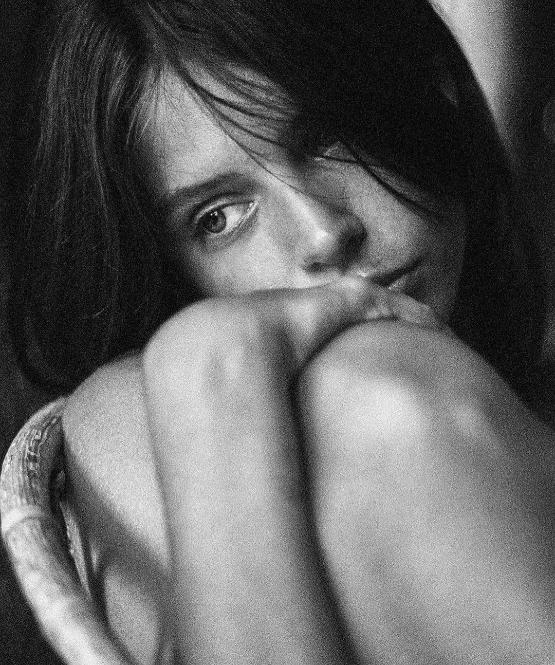 Black and white portrait of a young child with dark hair resting their chin on their knees while seated. Indoor setting, medium shot.