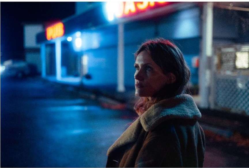 Woman in coat stands outside a brightly lit building at night, looking thoughtfully off-camera in a medium-shot portrait.