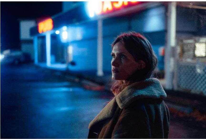 Woman in coat stands outside a brightly lit building at night, looking thoughtfully off-camera in a medium-shot portrait.