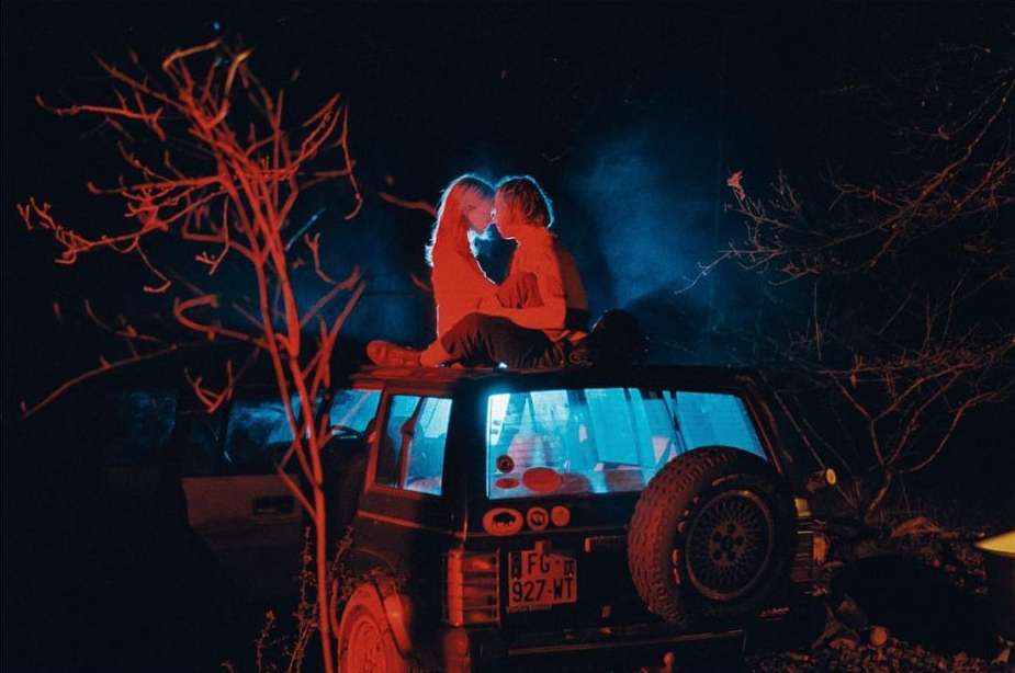 Two people sit on top of an SUV at night, illuminated by red and blue light, with trees visible in the foreground and background.