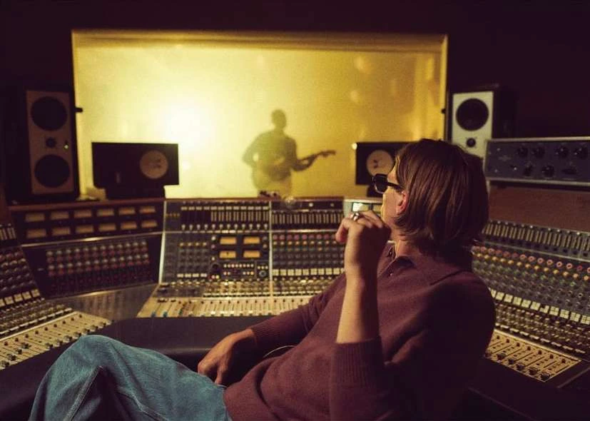 Man with sunglasses sits at sound mixing console in a music studio; a guitarist can be seen through glass in the background.