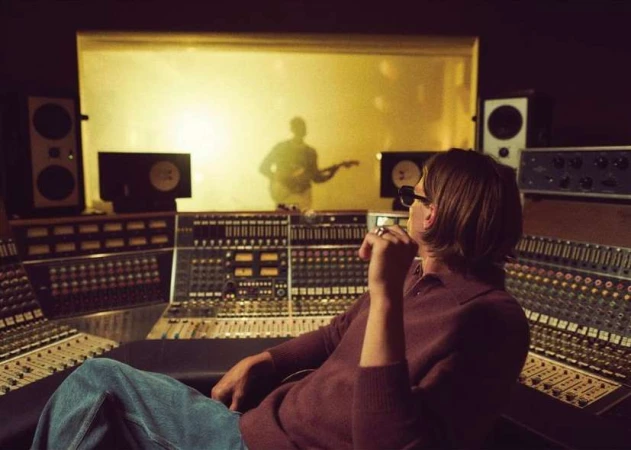 Man with sunglasses sits at sound mixing console in a music studio; a guitarist can be seen through glass in the background.