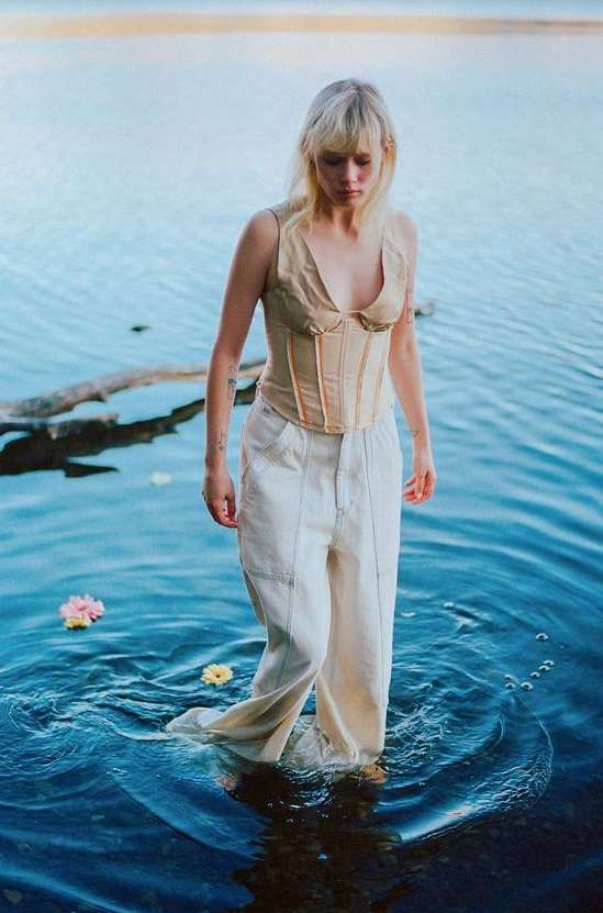 Blonde adult female in beige corset and white trousers wading in water with flowers, photographed at eye-level in an outdoor setting.