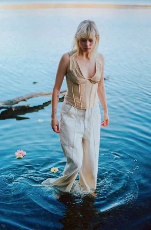 Blonde adult female in beige corset and white trousers wading in water with flowers, photographed at eye-level in an outdoor setting.