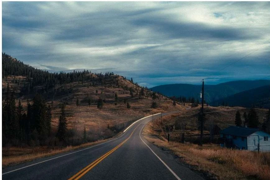 Scenic view of an asphalt road curving through a mountainous landscape under a cloudy sky, with sparse vegetation and distant mountains.