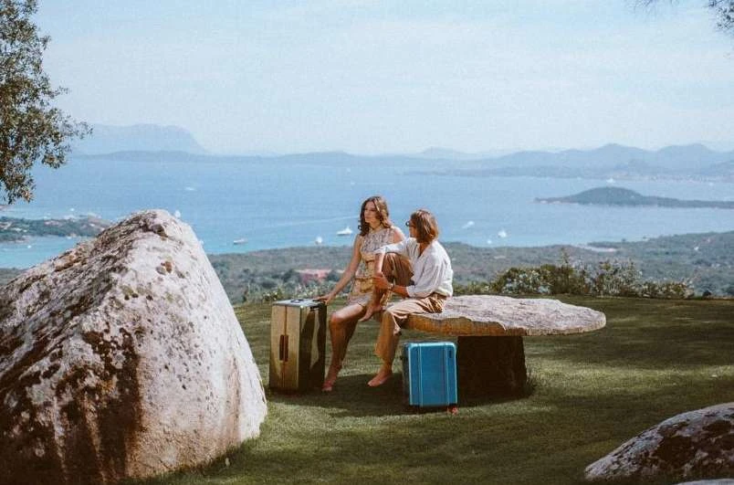 Couple sits on a stone bench overlooking the sea, with vintage luggage, enjoying a scenic view on a sunny day in a travel setting.