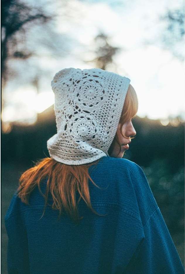 Woman with auburn hair wearing a crocheted bonnet and a blue denim jacket, photographed outdoors in soft, diffused lighting.