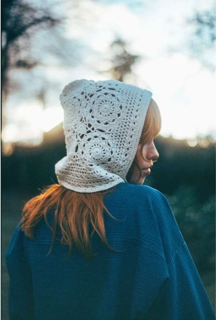 Woman with auburn hair wearing a crocheted bonnet and a blue denim jacket, photographed outdoors in soft, diffused lighting.