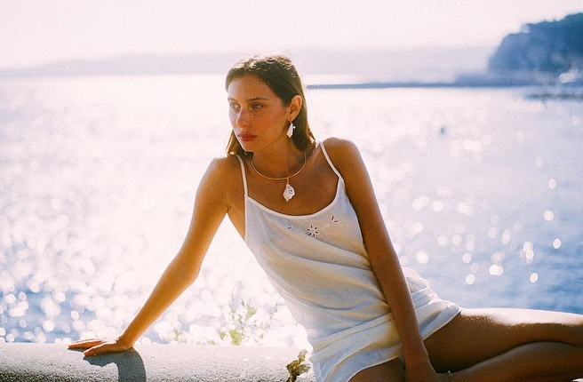 A young woman with long brown hair, wearing a dress and jewelry, is resting outdoors with the ocean and coastal hills in the background.