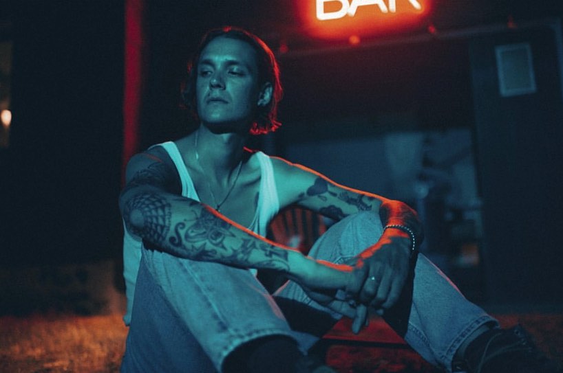 Tattooed man sitting outside a bar at night under the red neon sign, wearing a white tank top and blue jeans, looking off camera.